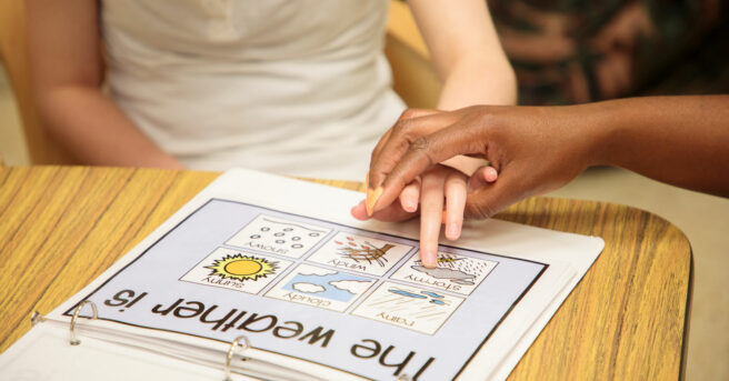 A student's supported hand using a communication binder with weather tiles. 
