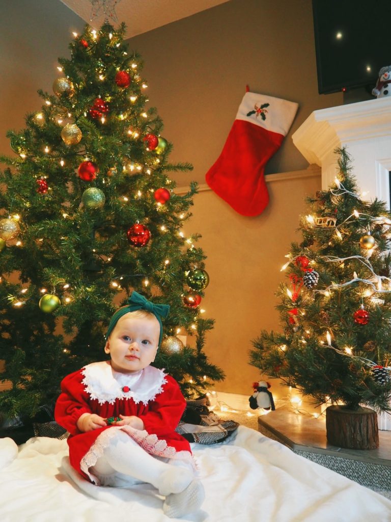 little girl sitting in front of Christmas tree wearing red dress, white stockings, and green bow in her hair. Attribute:  Shaylyn@mpadb