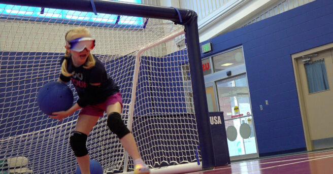 A girl wearing eye shades, knee pads ready to throw a goalball near the net.