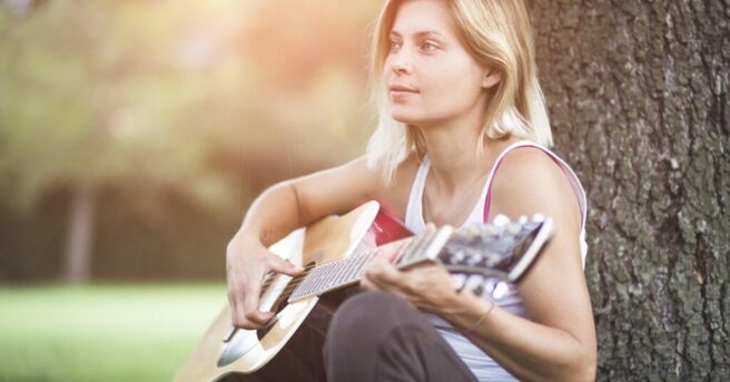 Young adult leans against a tree and strums guitar