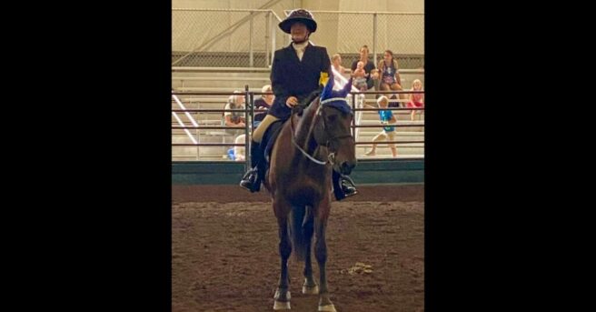 Leslie Weilbacher on a horse, Major, in the arena wearing a helmet with a large visor and a yellow armband on a black coat. The horse has his mane up in braids and is wearing a royal blue ear bonnet and saddle pad.