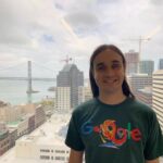 Lucas is standing straight to the camera, with a big smile in a friendly posture. Behind him is possible to see San Francisco skyline, bridge and sea. The sky is cloudy. Lucas is wearing a green T-shirt with Google’s logo that has a dragon placed as the second O and G of the spelling.