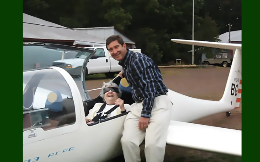 Jane Henkler is smiling and sitting inside a plane while her son, Ed, is also smiling and leaning on the wing.
