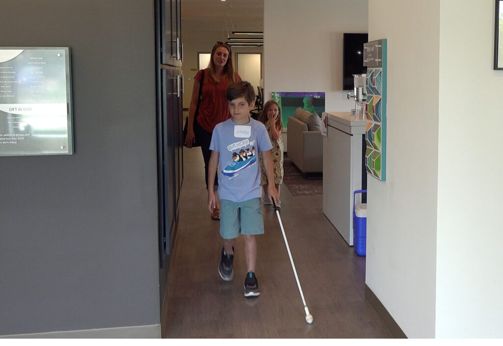 A student walking with his cane in an office area with his mom and sister.