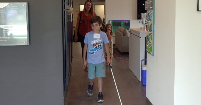 A student walking with his cane in an office area with his mom and sister.