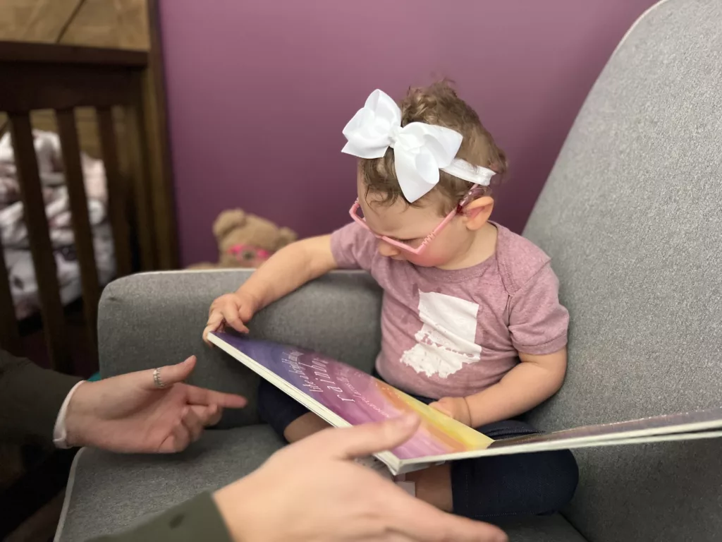 small child with pink eyeglasses sits in the corner of a large chair and explores a book with the help of a parent