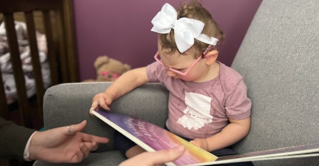 small child with pink eyeglasses sits in the corner of a large chair and explores a book with the help of a parent