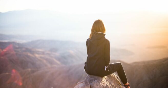 Young adult sits alone on a rock in a contemplative manner overlooking mountains