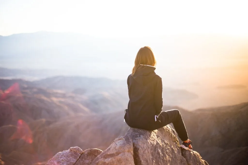Young adult sits alone on a rock in a contemplative manner overlooking mountains