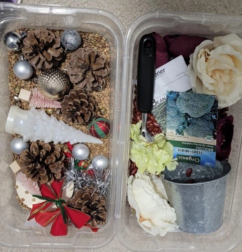 In the picture there are two sensory boxes. The one on the left is a Christmas sensory box, made of lentils, Christmas ornaments, a plastic light-up Christmas tree, Christmas bows, and a miniature Christmas tree. On the right, there is a spring sensory box made of beans, bags of seeds, a bucket, fake flowers, and a scooper.