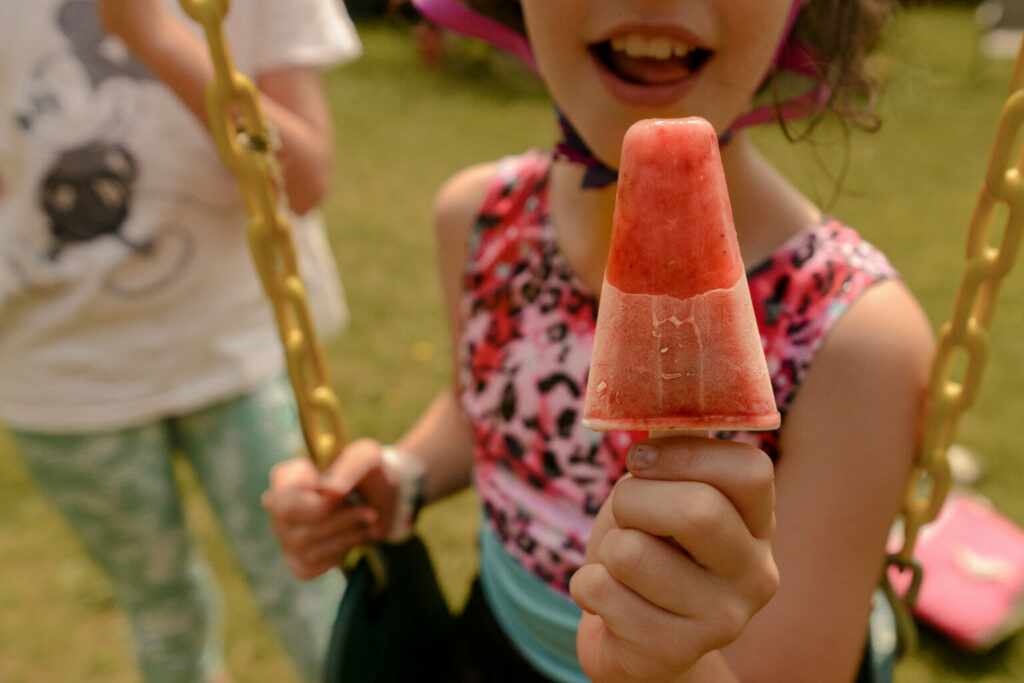 a smiling child is sitting on a swing and holding a popsicle out toward the camera