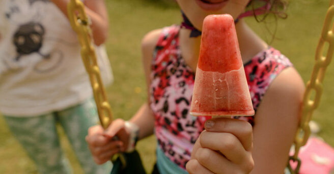 a smiling child is sitting on a swing and holding a popsicle out toward the camera