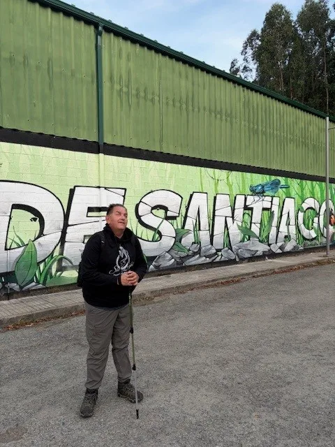 Richard Center stands with a hiking pole in front of a large ‘DE SANTIAGO’ graffiti mural.”