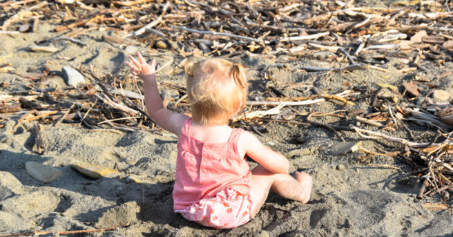 Back view of baby girl in pink with pony tails, waving her hand sitting in sandy beach.