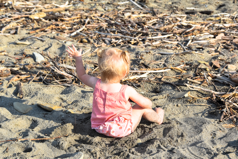 Back view of baby girl in pink with pony tails, waving her hand sitting in sandy beach.