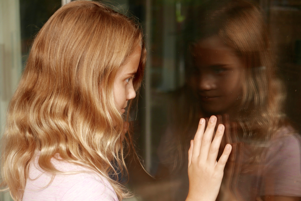 Young girl facing outside with her reflection of herself in the window. 