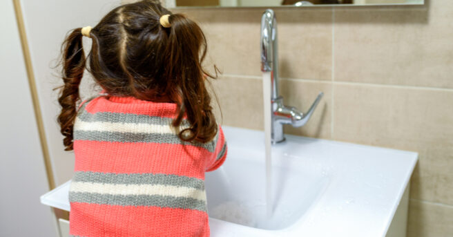 Back view of a girl with pig tails washing her hands and face in a bathroom. 