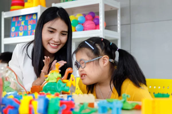 Student wearing glasses in a classroom setting sitting next to a teacher smiling and encouragingly clapping.