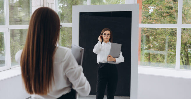 Individual wearing eyeglasses and professional clothing looks at her reflection in a mirror