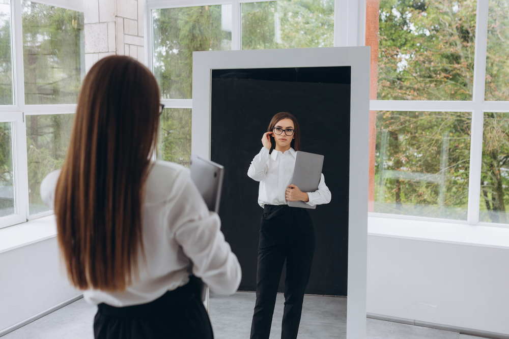 Individual wearing eyeglasses and professional clothing looks at her reflection in a mirror
