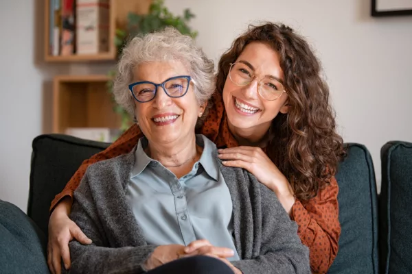 Older woman sitting on couch with younger woman behind her with arms around her. 