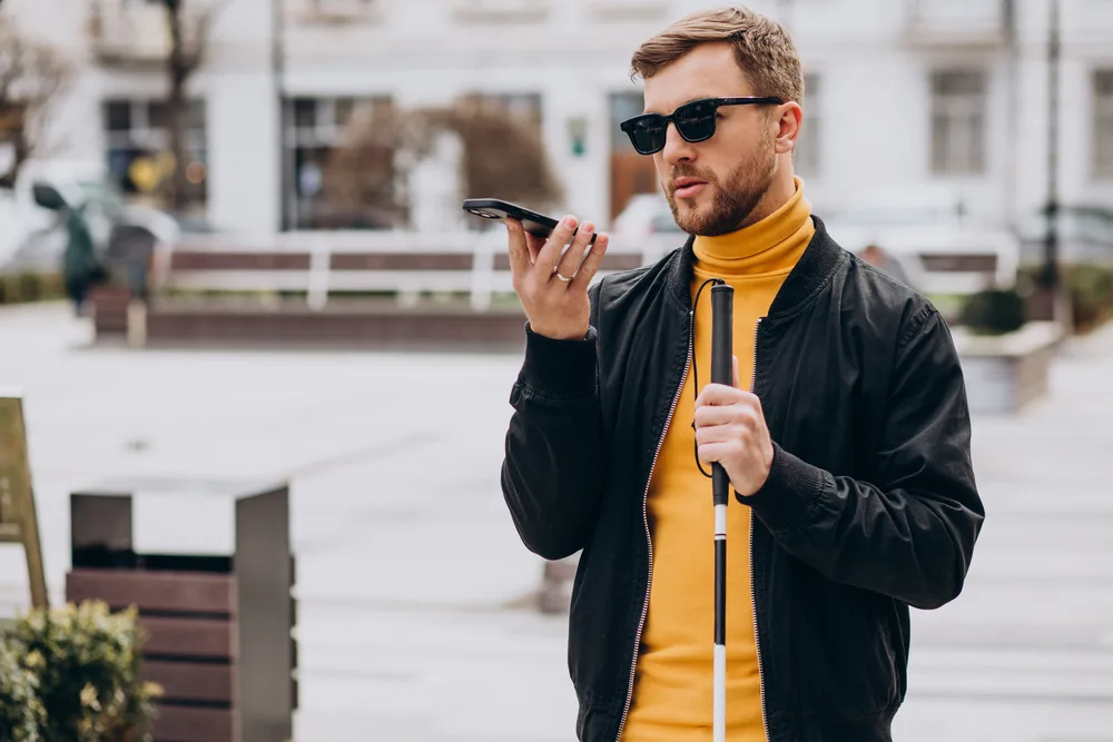 Young blind man with sunglasses holding a white cane outside talking into his phone.
