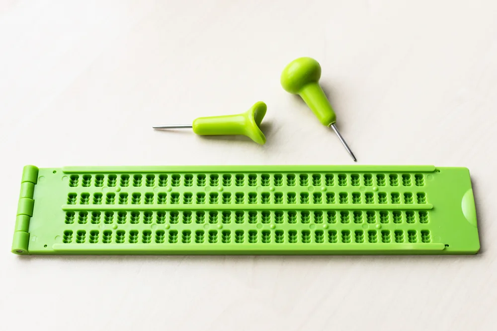 Plastic braille writing slate and stylus on brown table.