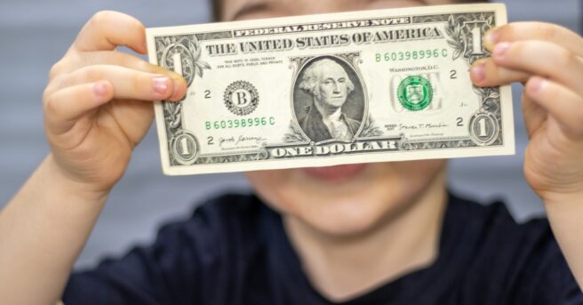 One dollar bill in front of kid child boy face cover eyes or mouth.