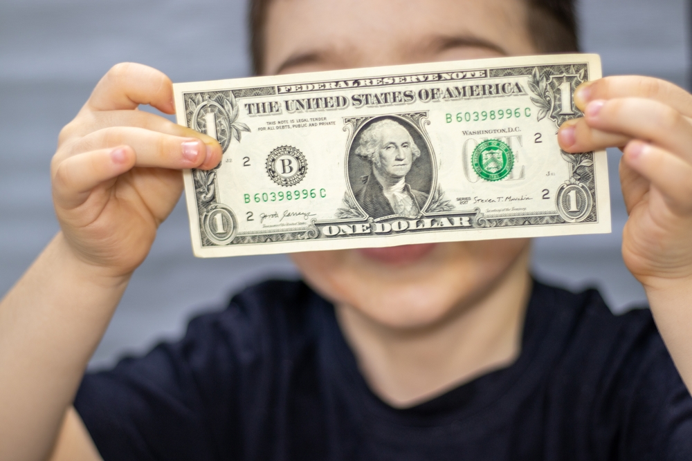 One dollar bill in front of kid child boy face cover eyes or mouth.