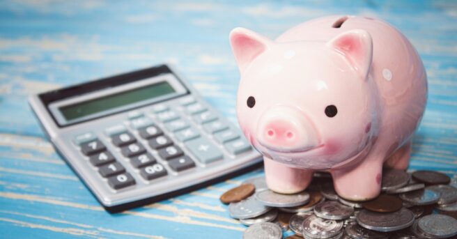 A piggy bank stands on a pile of coins next to the calculator.