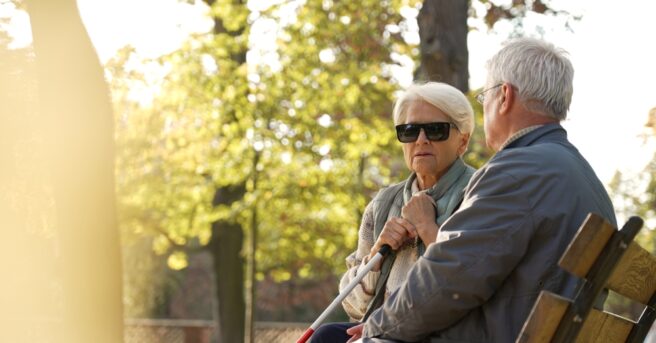 Older couple, one with a white cane, sitting on a bench