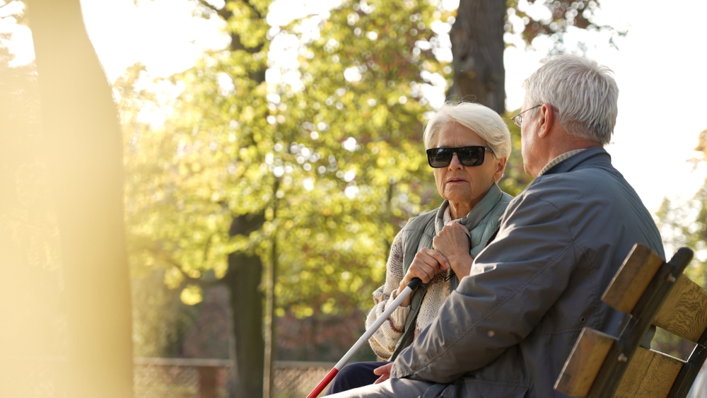 Older couple, one with a white cane, sitting on a bench