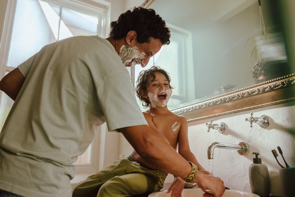 Adult shaves their face while nearby child pretends to do the same