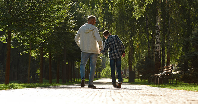 Back view of caucasian father and teenage son with cerebral palsy walking on pavement in sunny park.