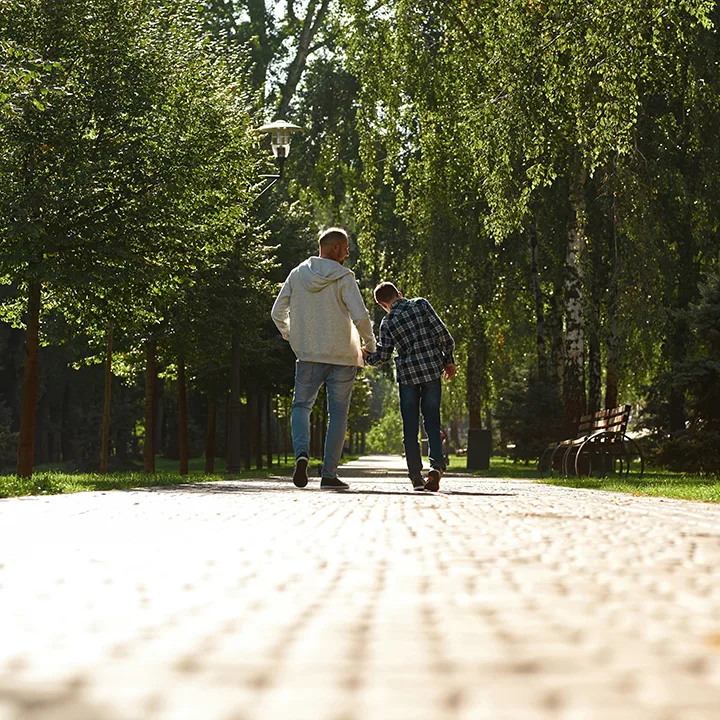 Back view of caucasian father and teenage son with cerebral palsy walking on pavement in sunny park.