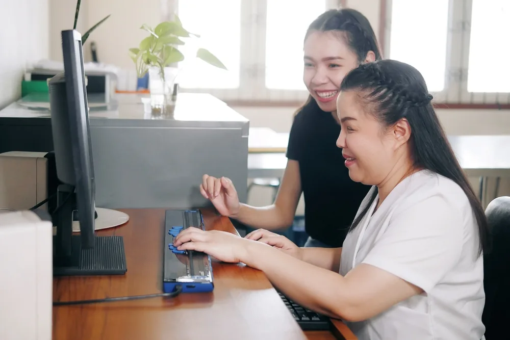 women co-workers in office workplace including person with blindness disability using computer with refreshable braille display assistive device.