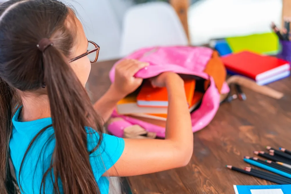 Cute happy schoolgirl puts stationery in a backpack.