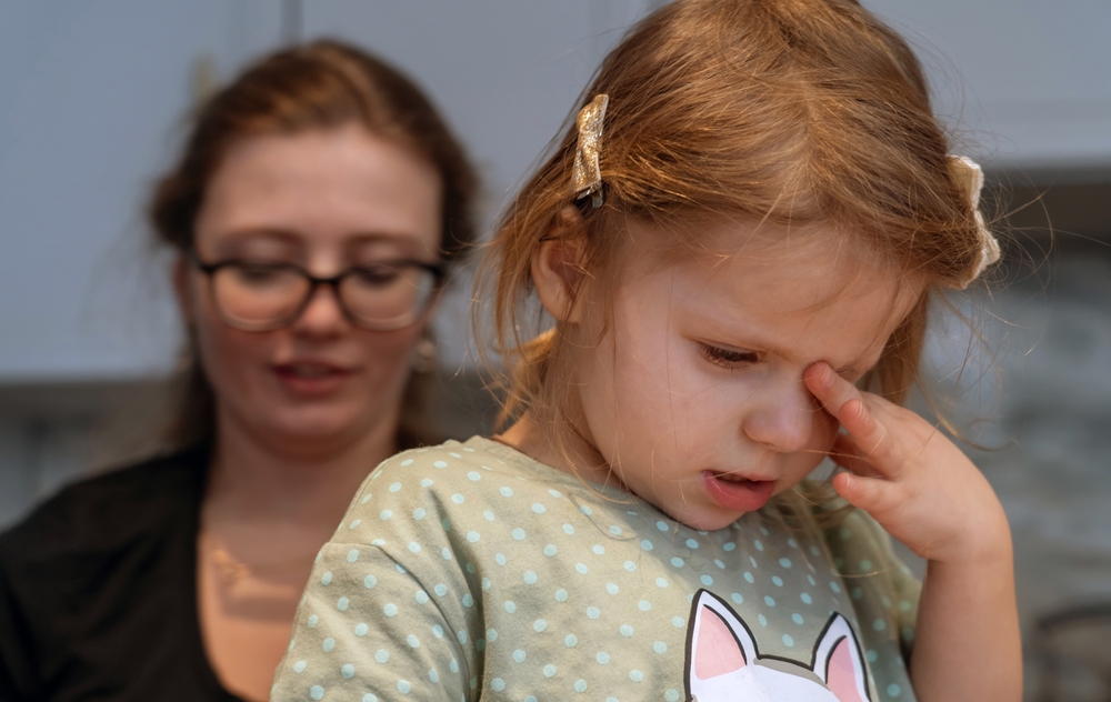 A little girl eye poking with her mom standing behind her. 