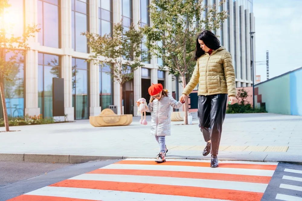 Young mother crossing street holding hand with daughter wearing eyeglasses looking down at crosswalk. 
