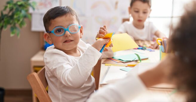 A preschooler wearing glasses and patch, sitting at a table cutting paper.