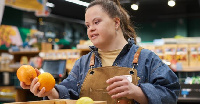 Waist up portrait of smiling young woman with disability working in supermarket sorting fresh fruits and produce