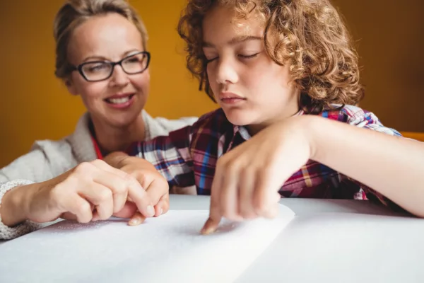 Boy using braille to read at school with teacher looking over his shoulder.