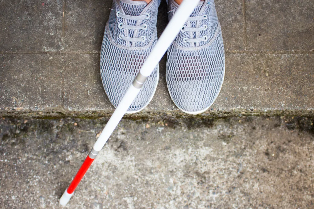 Top-down view of gray sneakers at a curb with a white cane with a red tip touching the edge of the step.