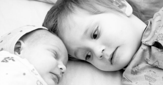 Toddler boy and his newborn sister are lying on a bed. Black and white photo.