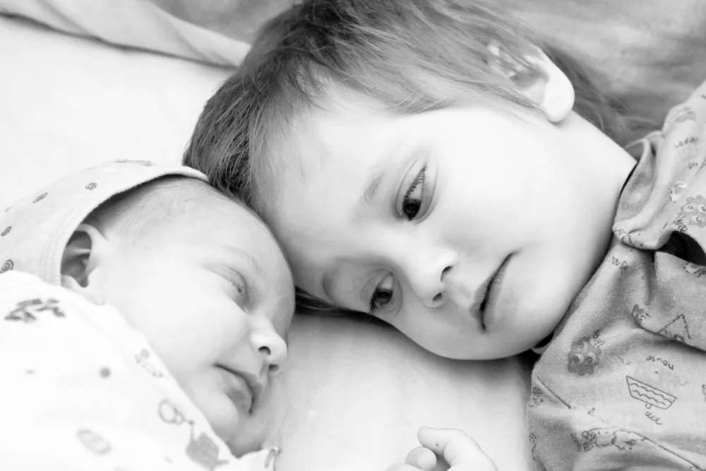 Toddler boy and his newborn sister are lying on a bed. Black and white photo.