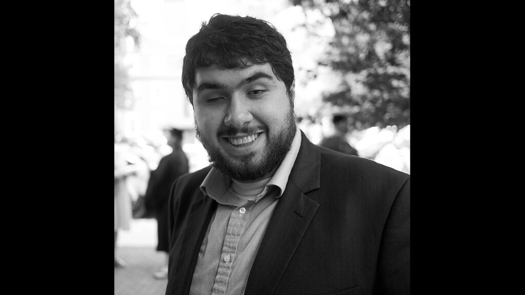 A black and white headshot of a Persian man with light skin and a toothy grin. Shown from the chest up, he has a full beard and thick, wavy dark hair. On our left, his right eye is closed, his thick arched eyebrows raised slightly. He wears a charcoal grey sports coat over a light-colored button-down shirt. The background is out of focus, with people in graduation gowns and fuzzy foliage.