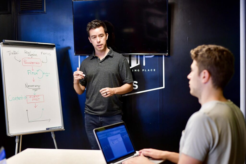 Coworkers in discussion; one is on a laptop and the other is at the whiteboard