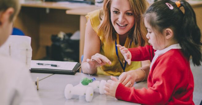 Teacher talks to young student in a red sweater who is touching a balloon-powered model car