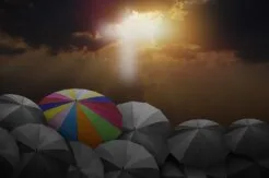 Umbrella in different colors among other black and white umbrellas as light shines through clouds.