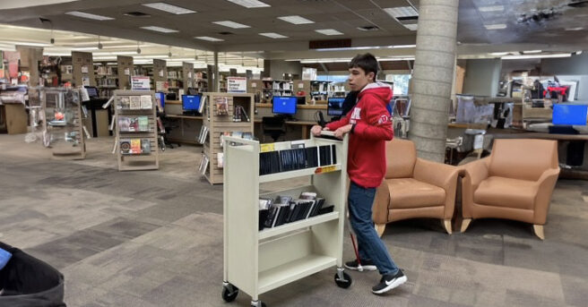 A teen pushing a cart with books in a library.